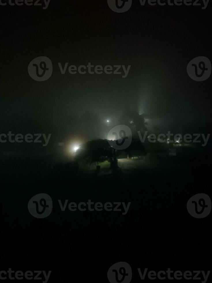 fir trees on meadow between hillsides with conifer forest in fog under the blue sky at night. spooky countryside scenery in full moon light photo