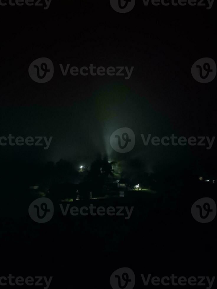 fir trees on meadow between hillsides with conifer forest in fog under the blue sky at night. spooky countryside scenery in full moon light photo