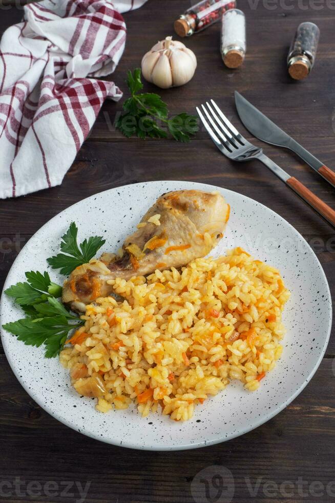 Delicious Asian pilaf, stewed rice with vegetables and chicken drumstick on a plate. Wooden rustic background. photo