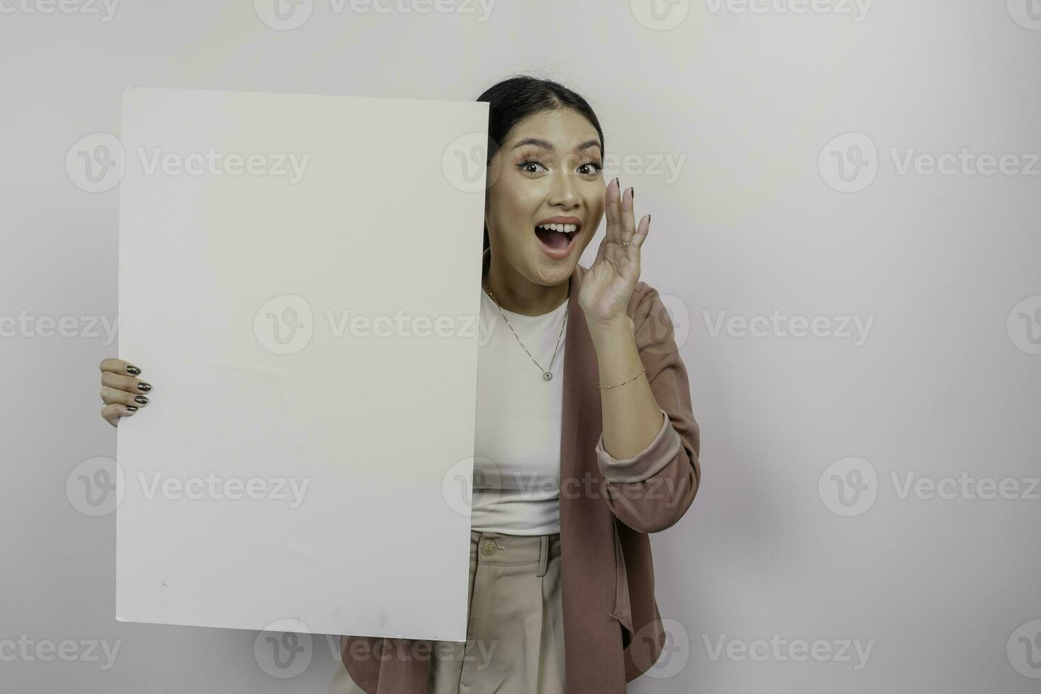 Young beautiful Asian woman employee wearing cardigan is shouting and screaming loud with a hand on her mouth while holding empty blank board, isolated by white background. photo