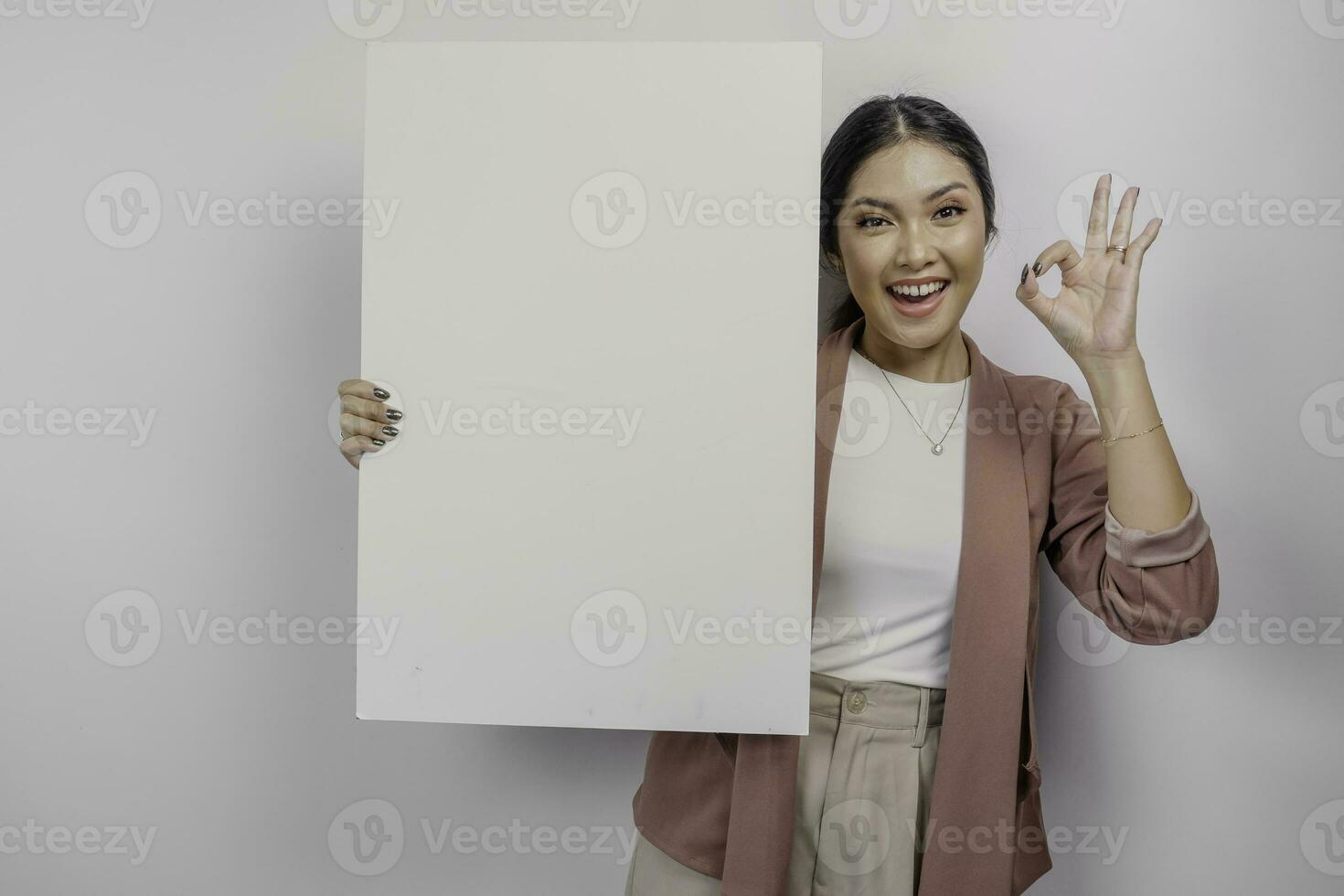 Smiling young Asian woman employee holding and showing empty blank board and gesturing OK sign with her fingers, isolated by white background photo
