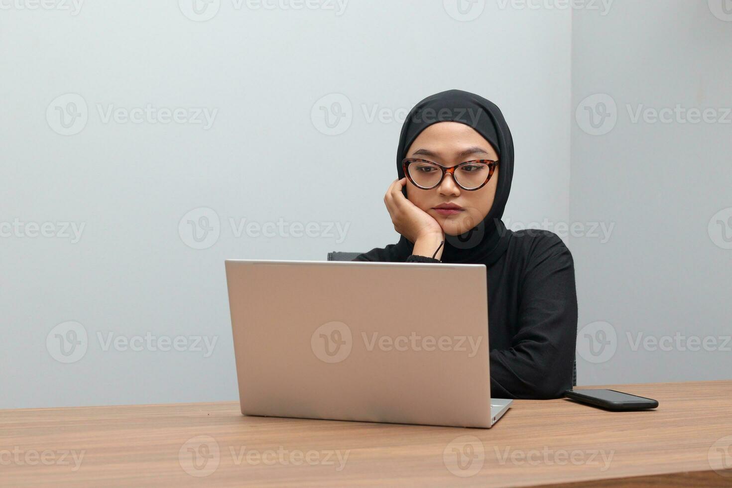 Portrait of attractive Asian hijab woman working on her laptop. Muslim girl feeling exhausted and tired. Employee and freelance worker concept. photo