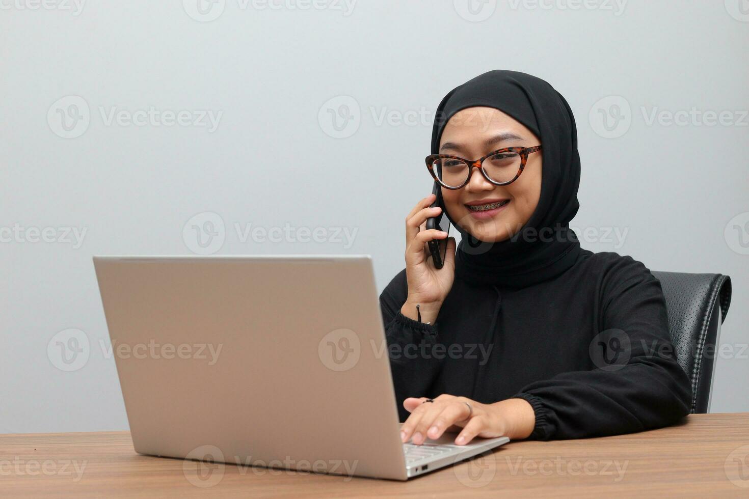 Portrait of attractive Asian hijab woman working on her laptop. Muslim girl making a phone call in office. Employee and freelance worker concept. photo