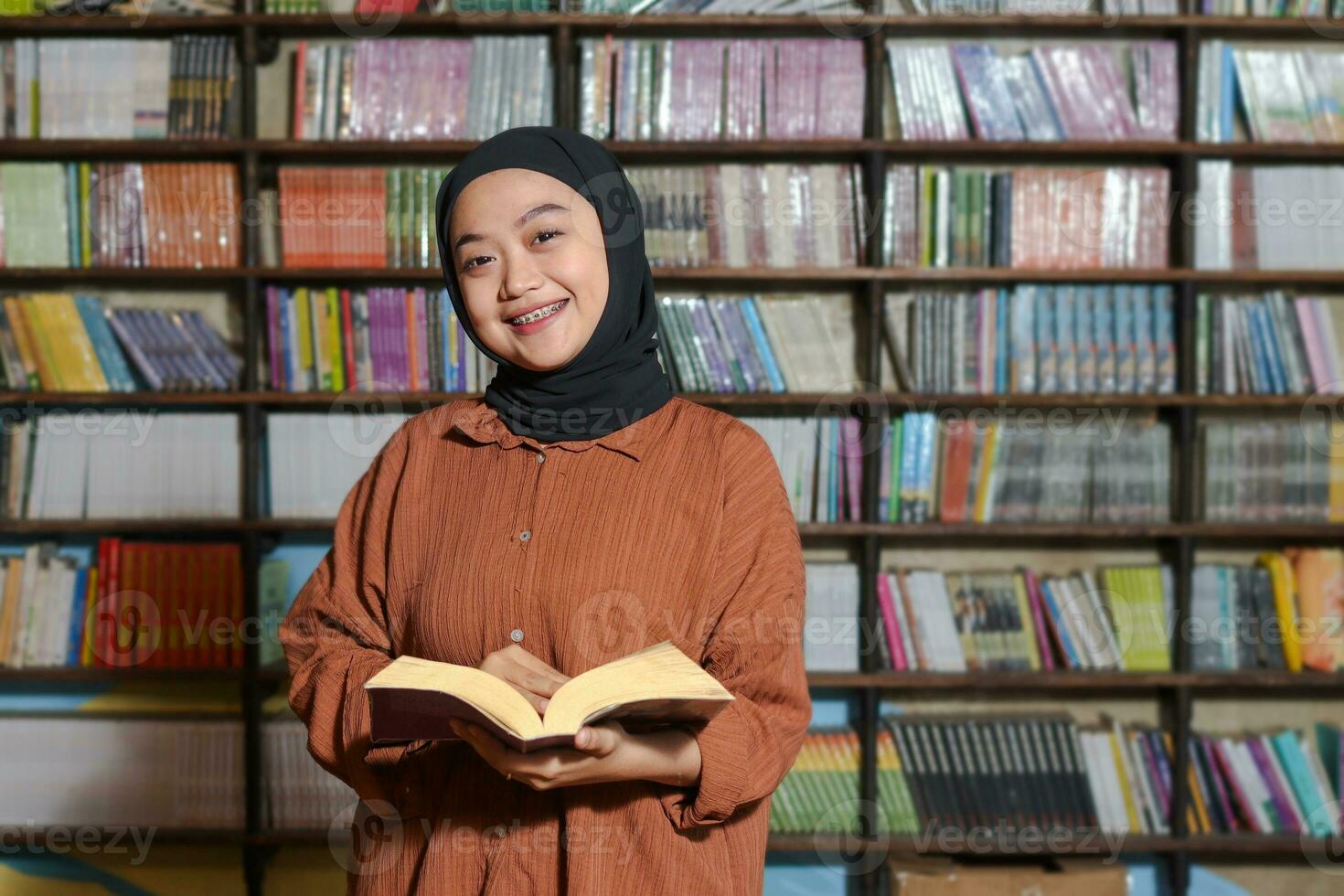 retrato de asiático hijab mujer participación libro en frente de biblioteca estante para libros. musulmán niña leyendo un libro. concepto de literatura y conocimiento foto