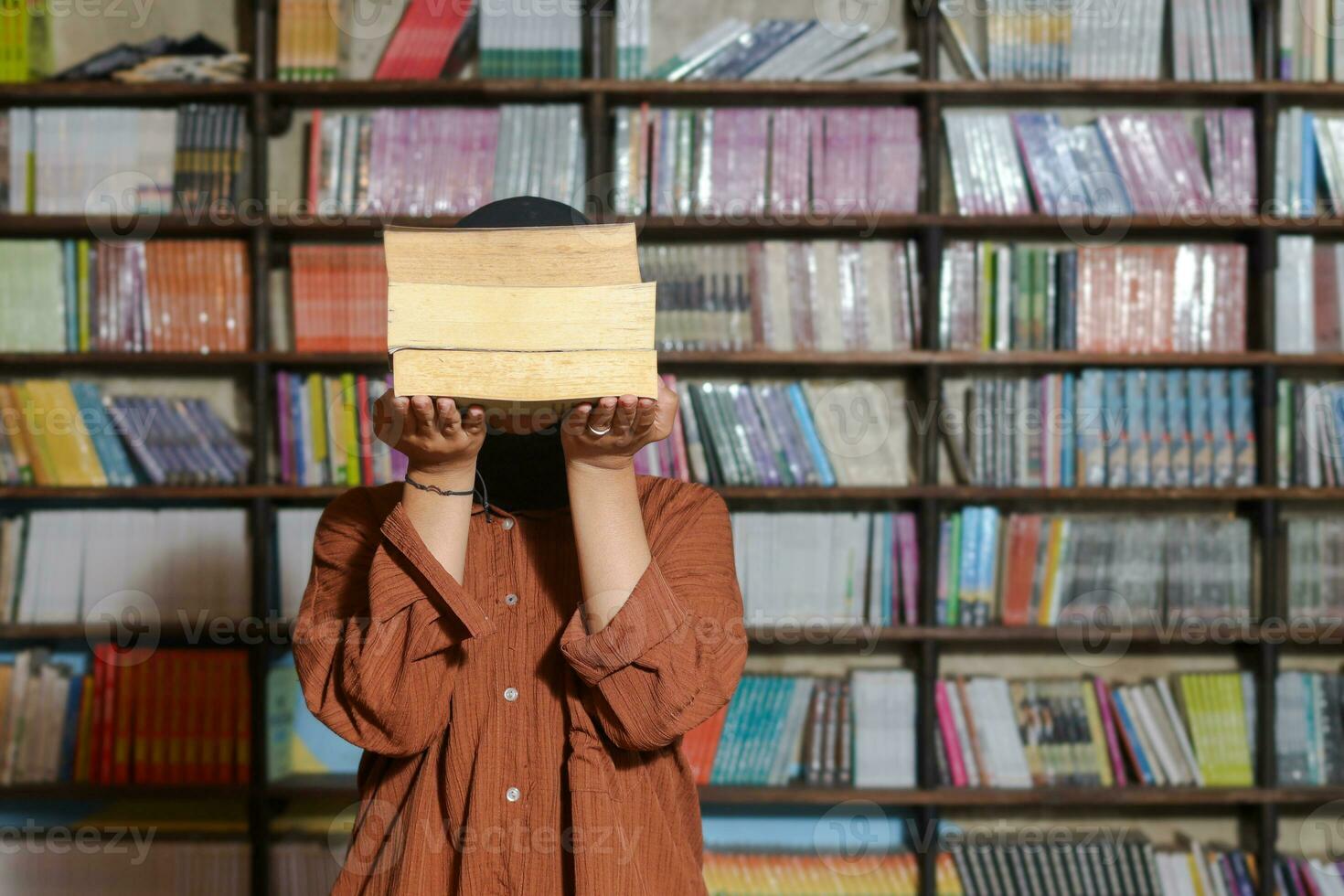 Portrait of Asian hijab woman covering her head with a pile of books in front of library bookshelf. Muslim girl reading a book. Concept of literacy and knowledge photo