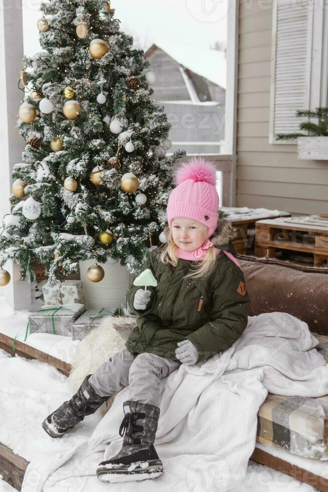 A little girl in winter clothes is sitting on the porch of a house decorated for the Christmas holidays. Christmas holidays for a child. photo