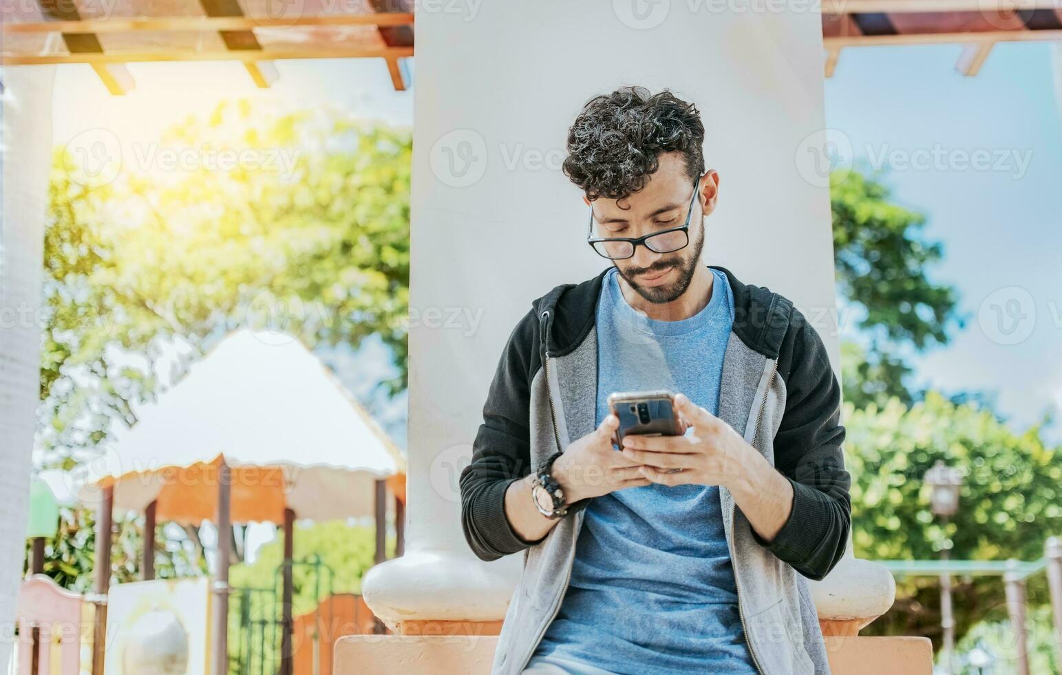 Handsome guy in glasses leaning on a wall chatting on his cell phone, Teenage male leaning on a wall sending a text message, Smiling handsome man using a phone leaning on a wall in a park photo