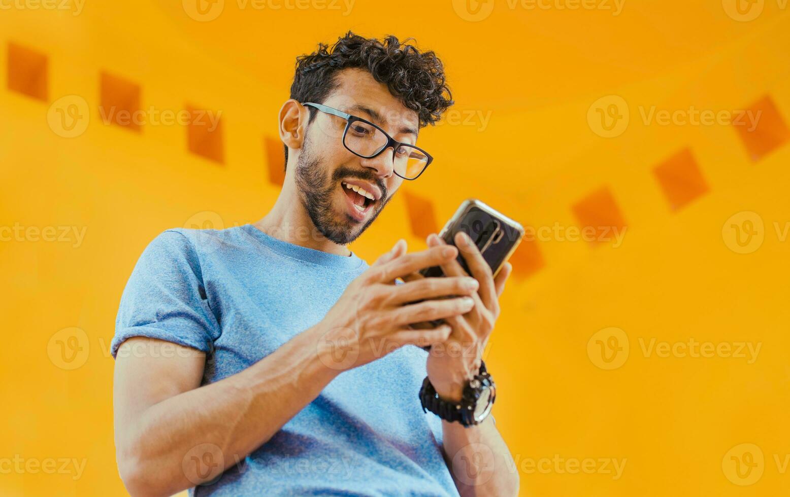 Happy guy sending a message with a cell phone near a yellow wall outdoors. Lifestyle of handsome bearded man using cell phone outdoors, Low angle view of young man using cell phone outdoors photo