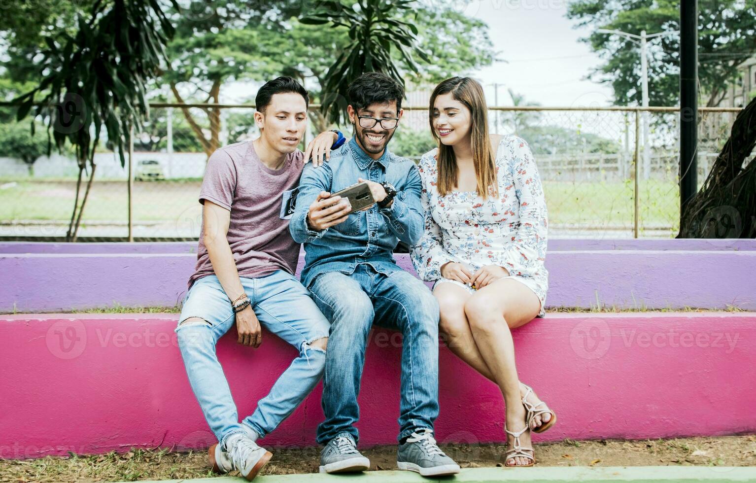 Tres contento amigos mirando a el célula teléfono sentado en el parque. Tres alegre personas mirando a célula teléfono sentado afuera, contento Adolescente amigos mirando a teléfono pantalla al aire libre foto