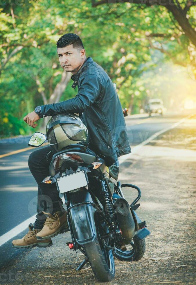 Portrait of biker in jacket sitting on his motorcycle at the side of the road. Latin motorcyclist sitting on his motorbike near a road photo