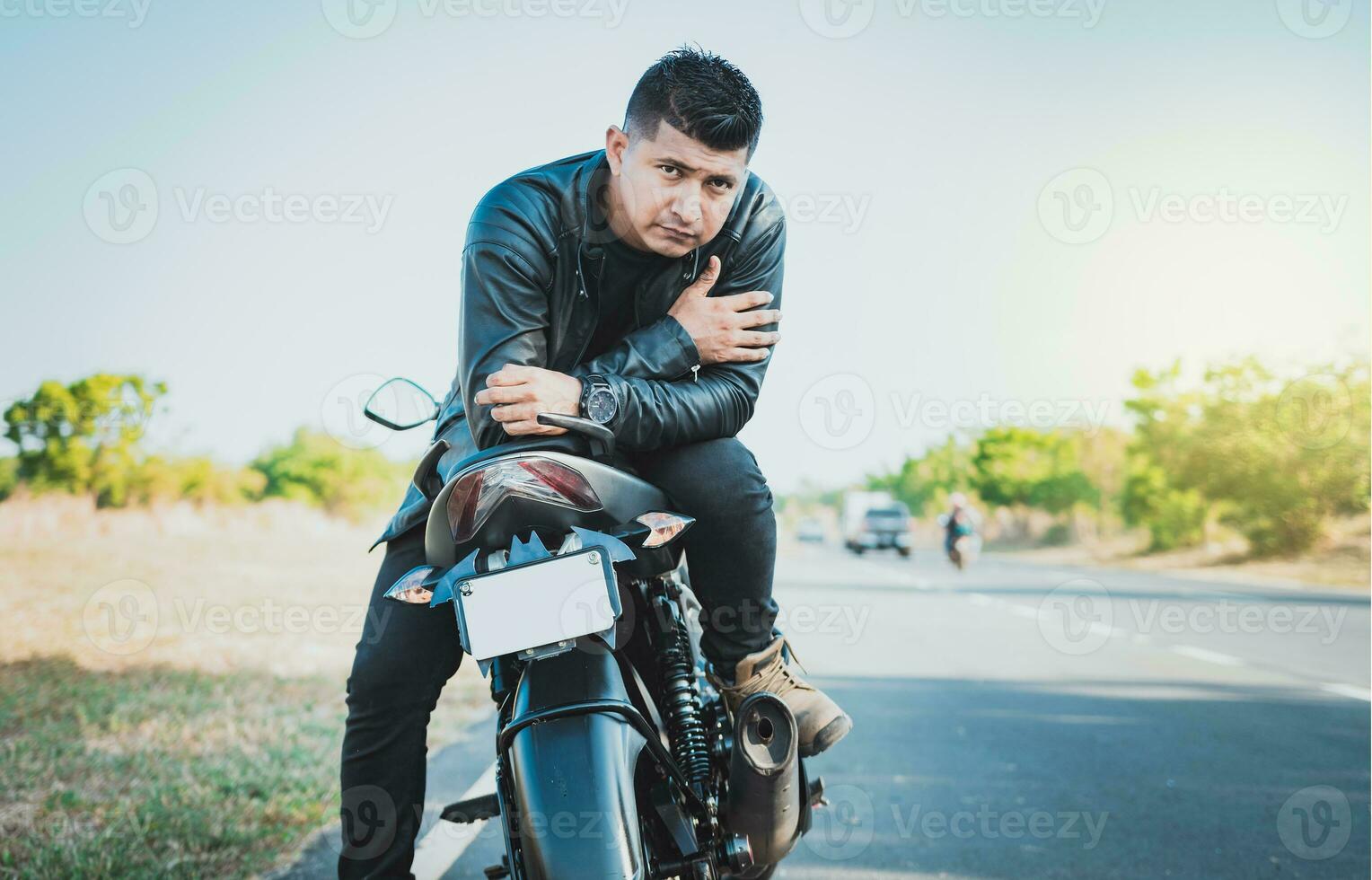 Portrait of handsome biker on his motorbike looking at camera outdoors. Handsome motorcyclist in jacket sitting on his motorcycle at the side of the road photo