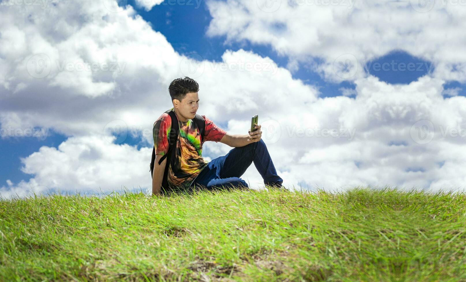 backpacker with phone on top of a hill , a man sitting on the grass with his cell phone photo