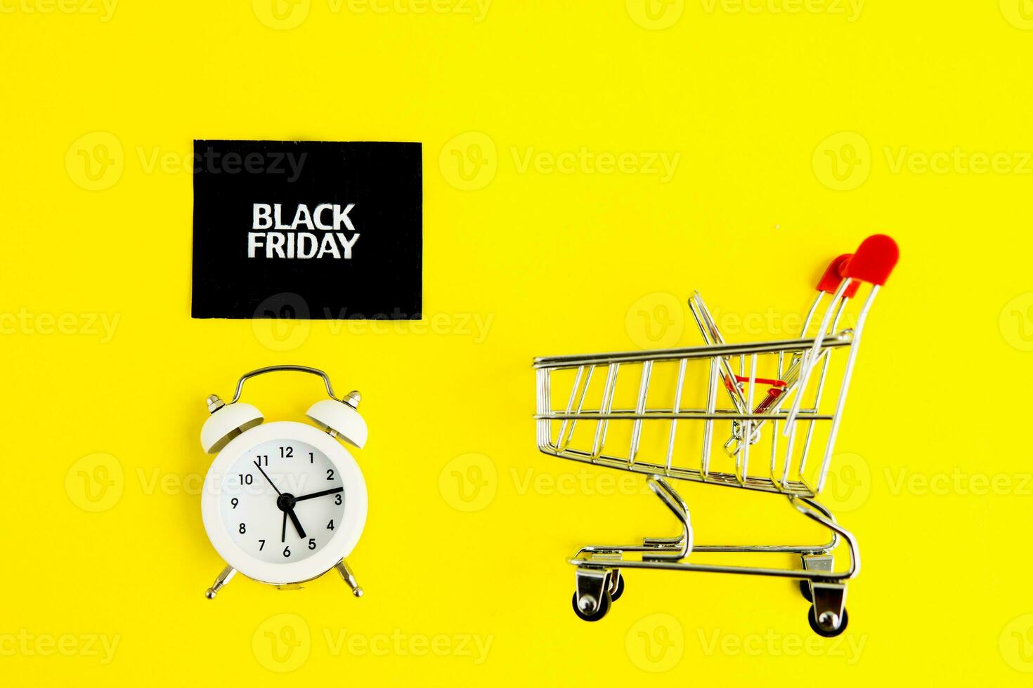 A white alarm clock, a shopping cart, and a black square that says black Friday on a yellow background. The topic of sales. photo