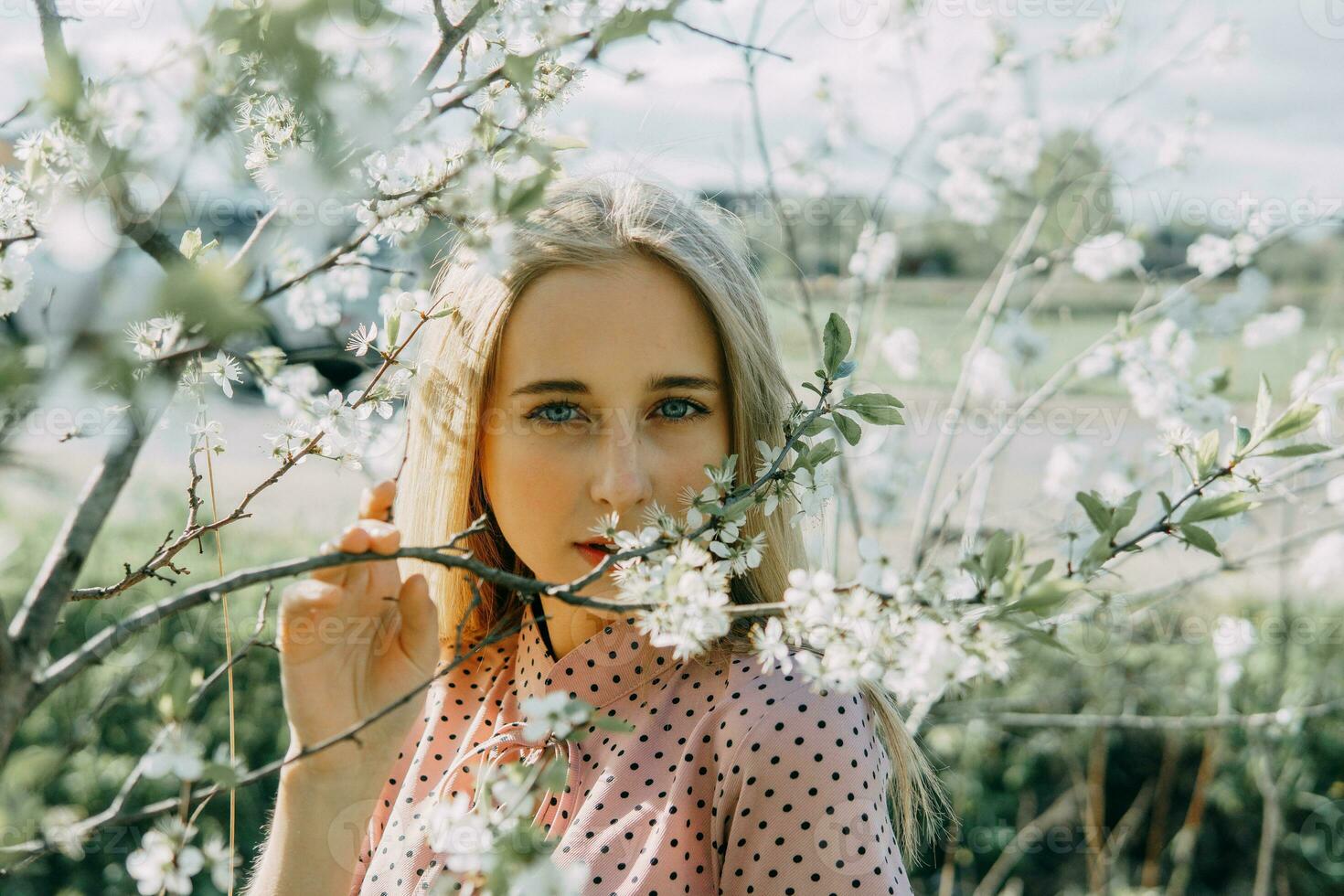 Blonde girl on a spring walk in the garden with cherry blossoms. Female portrait, close-up. A girl in a pink polka dot dress. photo