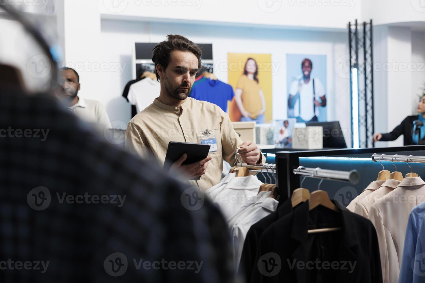 Clothing store young caucasian man manager using digital tablet to organize merchandise. Shopping mall employee holding portable gadget and hanging shirts on rack while working in boutique photo
