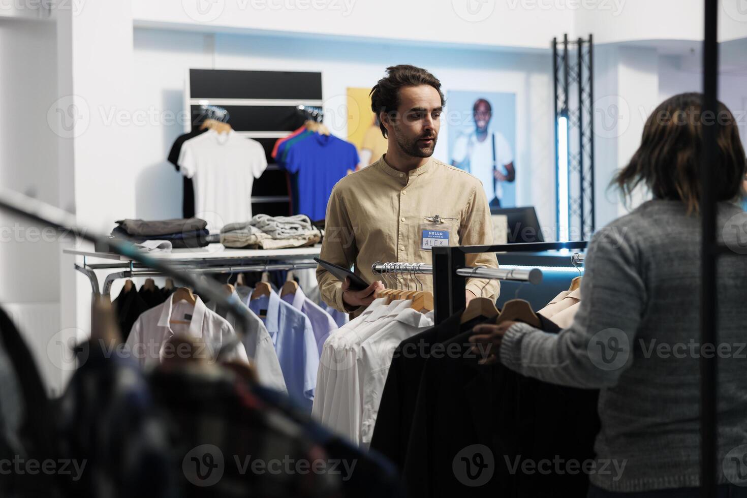 compras centrar empleado utilizando tableta a gestionar vestir valores mientras hablando con compañero de trabajo ropa Tienda caucásico hombre trabajador examinando prenda en sitio web mientras trabajando en boutique foto