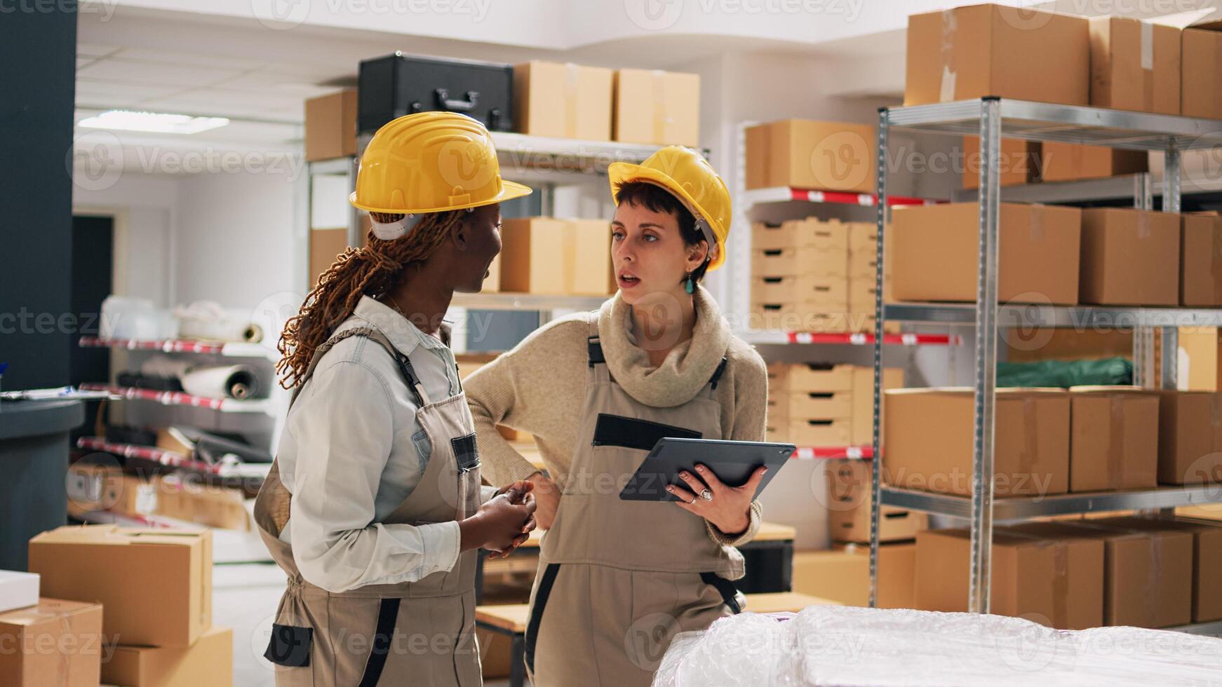 Two women with hardhats organizing products on shelves, looking at tablet to check stock logistics and planning distribution. Warehouse manager and employee counting cardboard packs. photo