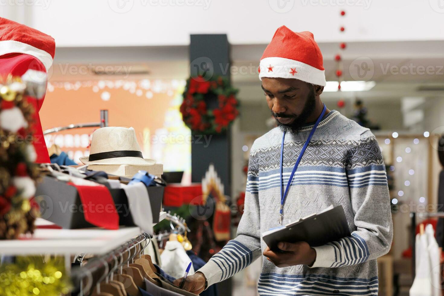 African american man counting hangers in clothing store, working on stock inventory while he wears santa hat. Retail sales assistant checking list of merchandise in shopping center mall. photo