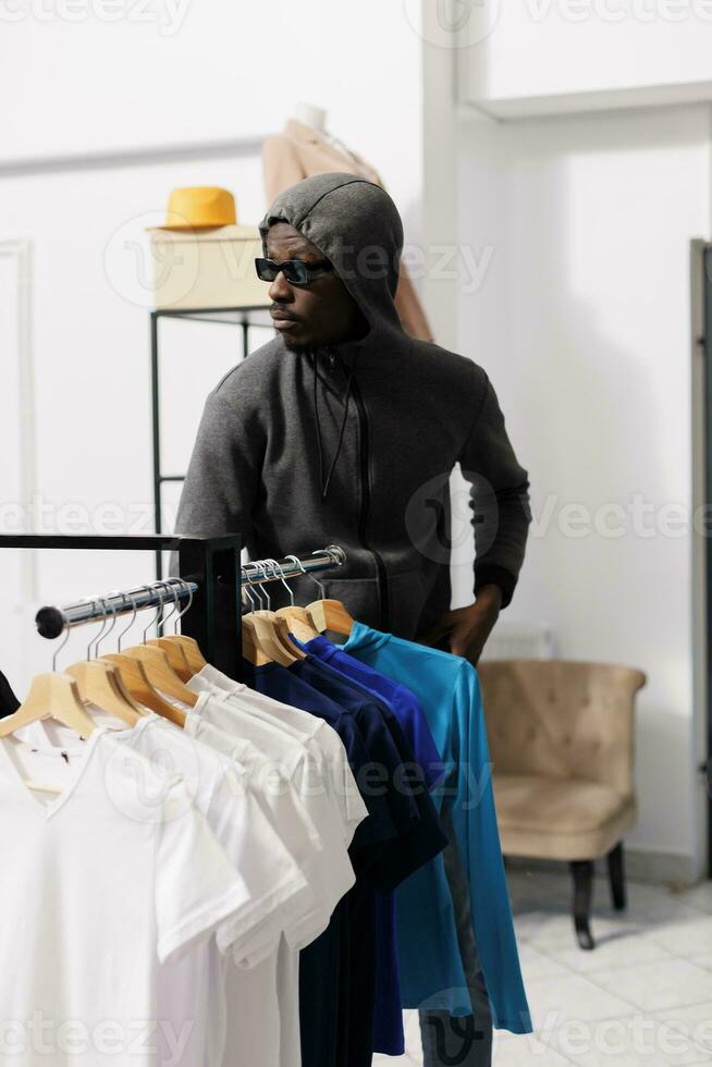 African american robber choosing to steal fashionable merchandise, looking around to see if someone watching him. Thief robbing modern boutique, wearing hood and sunglasses. Crime concept photo