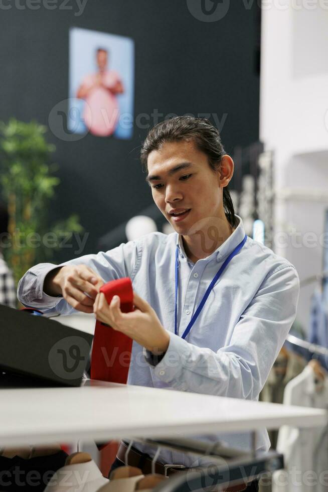 asiático hombre mirando a de moda accesorios, arreglando cinturón en Tienda estante en moderno boutique. elegante empleado trabajando en compras centro, trabajando con perchas lleno con ventas ropa foto