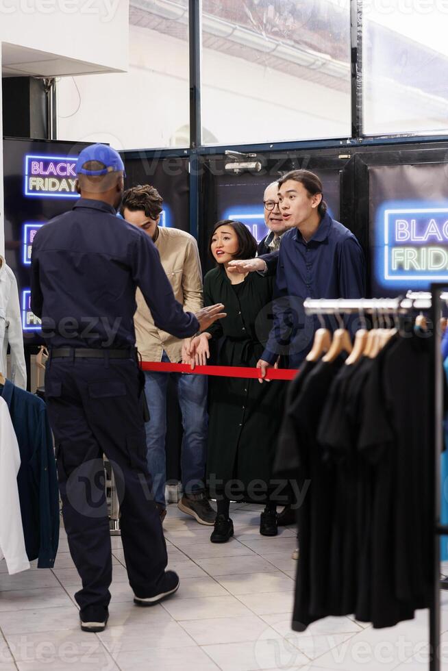 Security guard keeping waiting shopper crowd in check before stores open on Black Friday. Angry diverse people buyers standing behind control barrier with redtape in shopping mall during sales photo