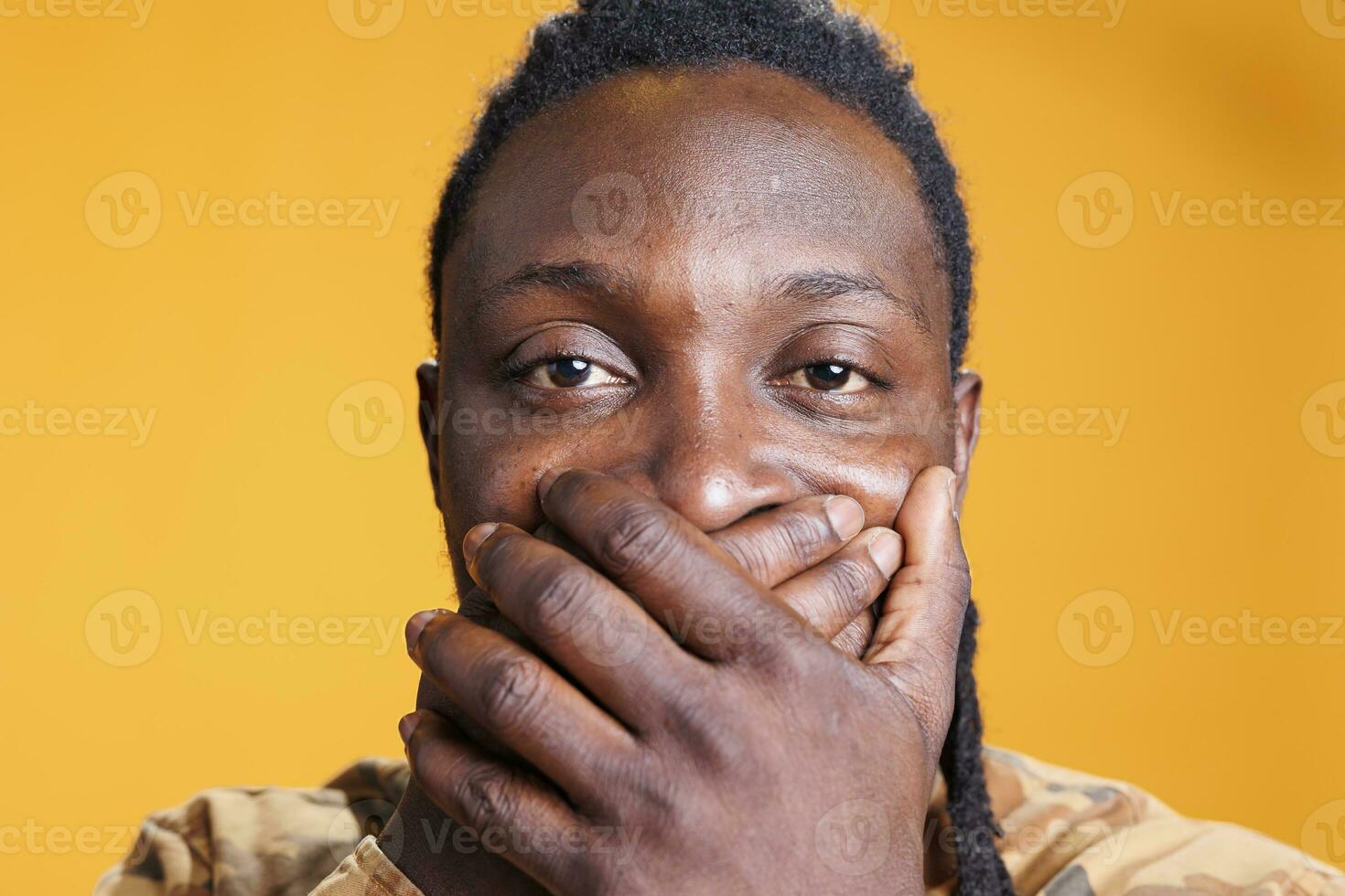 Serious man covering mouth with hands, showing speak no evil sign in studio over yellow background. African american young adult doing three wise monkeys gesture. Keeping secret concept photo