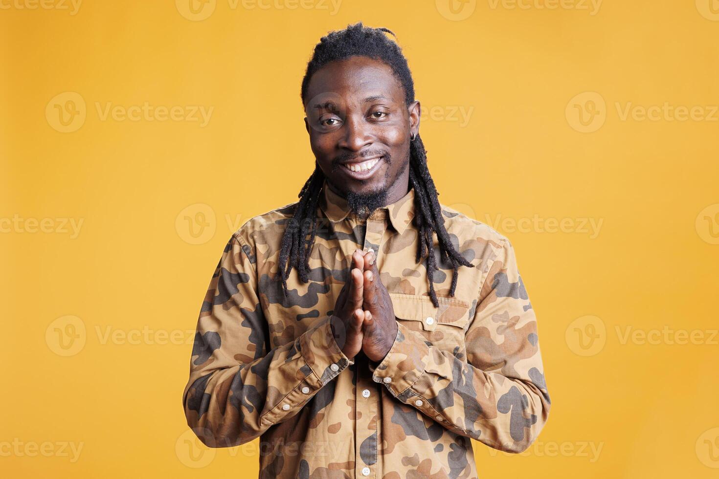Happy african american person holding palms together, asking for something in studio over yellow background. Smiling man celebrating succes, applauding people after work achievement photo