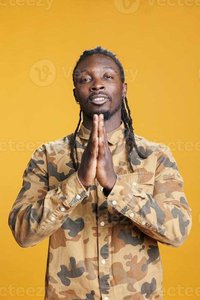 Portrait of peaceful man praying to god asking for forgiveness while standing in studio with yellow background. African american religious person holding palms together in pray. Religion concept photo