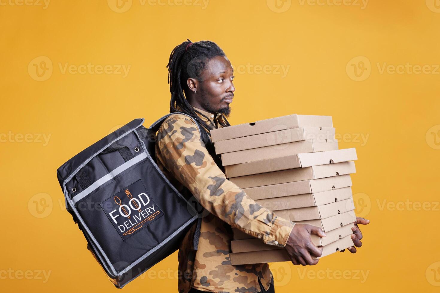 Takeout delivery worker holding stack with pizza cardboxes during takeaway delivering, standing in studio over yellow background. Smiling restaurant courier carrying thermal backpack. Food service photo