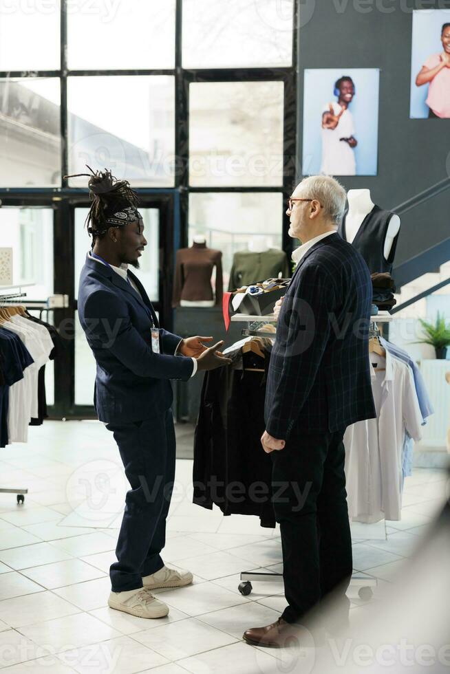 African american worker helping client with formal suit in clothing store, customer looking to buy modern clothes. Elderly client buying fashionable merchandise and trendy accessories in showroom photo