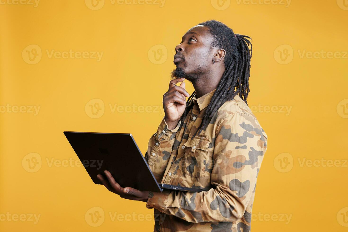African american man searching webpages on internet using laptop computer, standing in studio over yellow background. Young adult using portable gadget to navigate on social media photo