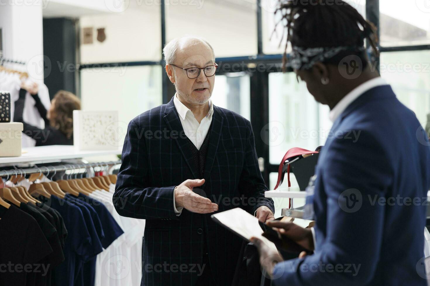 Elderly man asking worker for help with formal wear, african american employee checking merchandise inventory on tablet computer in showrrom. Senior customer looking at new fashion collection photo