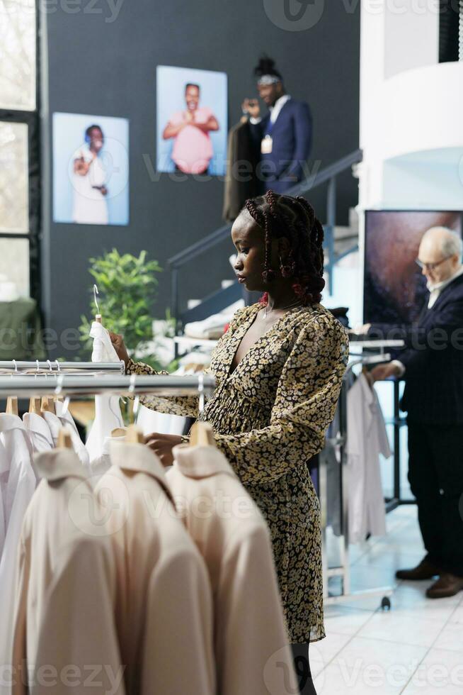 Pregnant woman looking at white shirt in modern boutique, shopping for fashionable maternity clothes. African american customer buying stylish casual wear in clothing store. Fashion concept photo