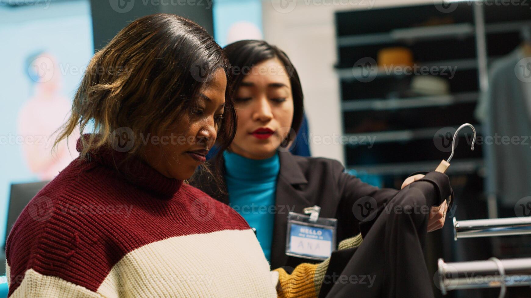 Female worker showing shirts to woman in boutique and buying trendy merchandise, talking about new clothes collection. Shop assistant working in clothing store, customer shopper. Handheld shot. photo