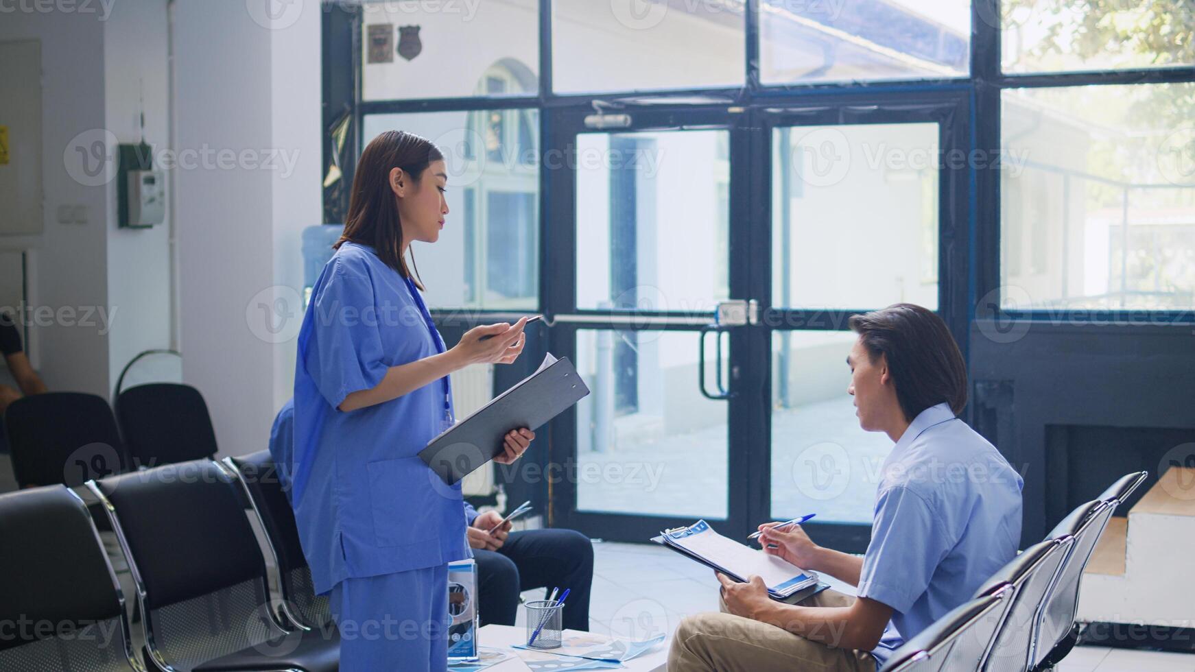 Assistant standing at hospital counter desk, writing medical expertise on clipboard. Asian patient calls the nurse to help him with insurance information during checkup visit in waiting room photo