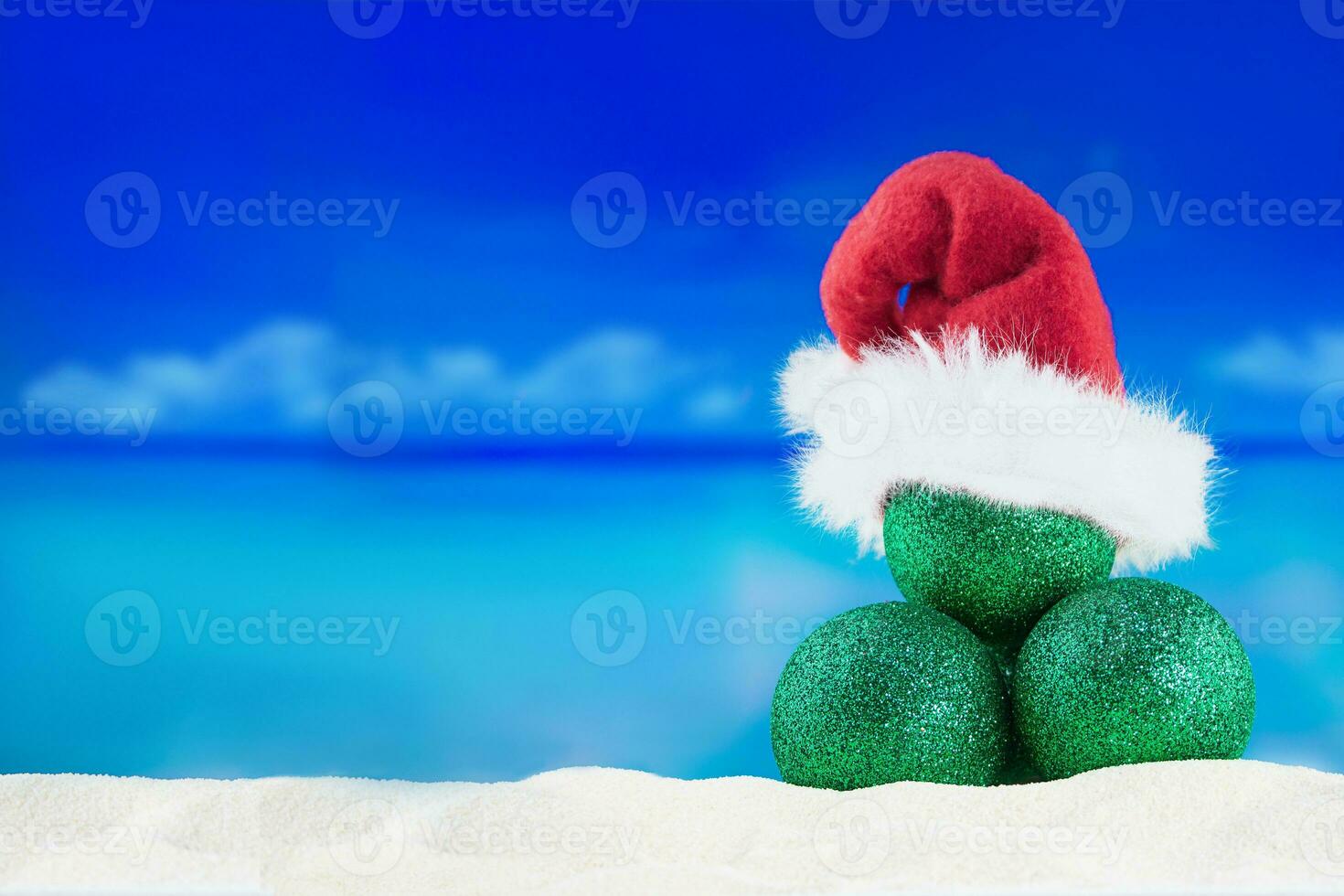 Shiny green balls in red Santa Claus' hat on sand of beach with sea. Christmas, New Year in hot countries. Copy space photo