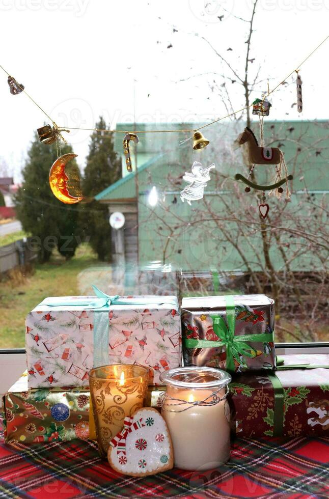 Christmas gifts, candles in glass in candlesticks with gingerbread in the shape of a heart near a window with a garland in a rural house, vertical photo