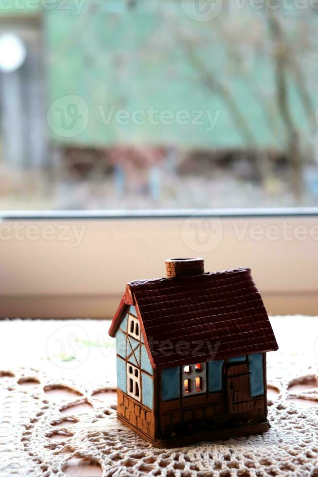 Ceramic candlestick in the shape of a house with a burning candle inside against a window in the village. Model of a house with burning windows, vertical photo, background with space for text photo
