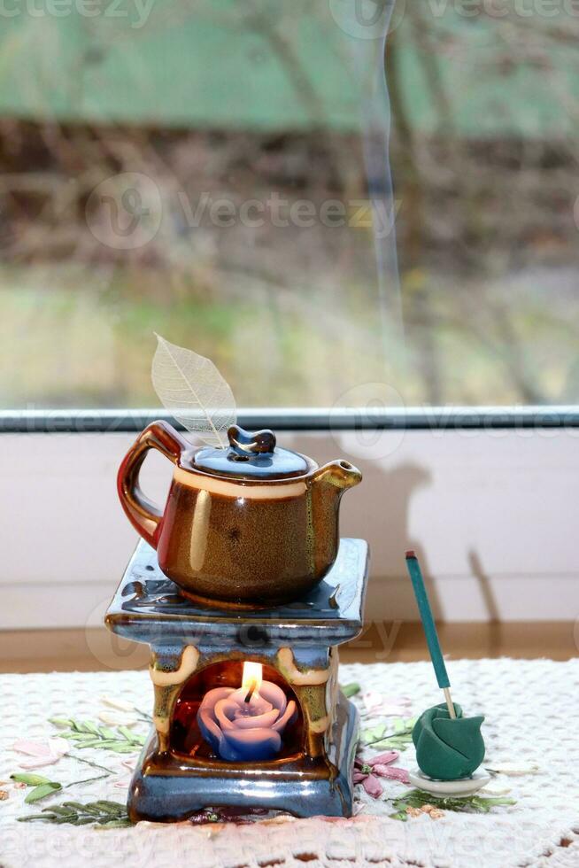 Aroma lamp in the form of a teapot with a burning candle inside and an openwork leaf and a burning incense stick by the window, vertical photo