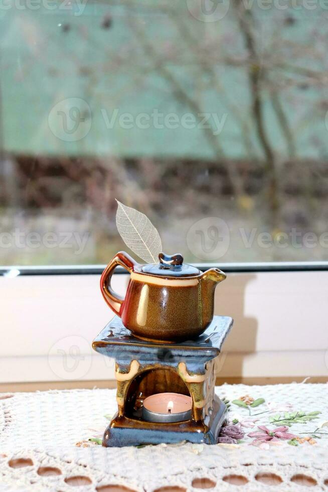 Aroma lamp in the form of a teapot with a burning candle inside near the window on a napkin, vertical photo