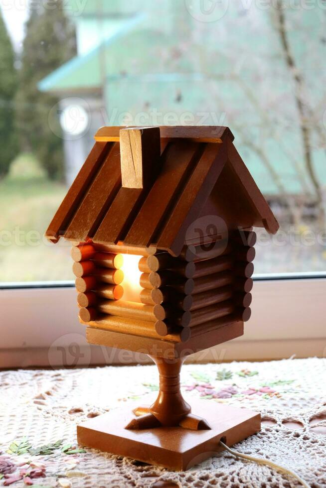 A wooden retro lamp in the form of a hut on chicken legs, a fairy-tale house with a burning window in the village. Vertical photo, close-up photo