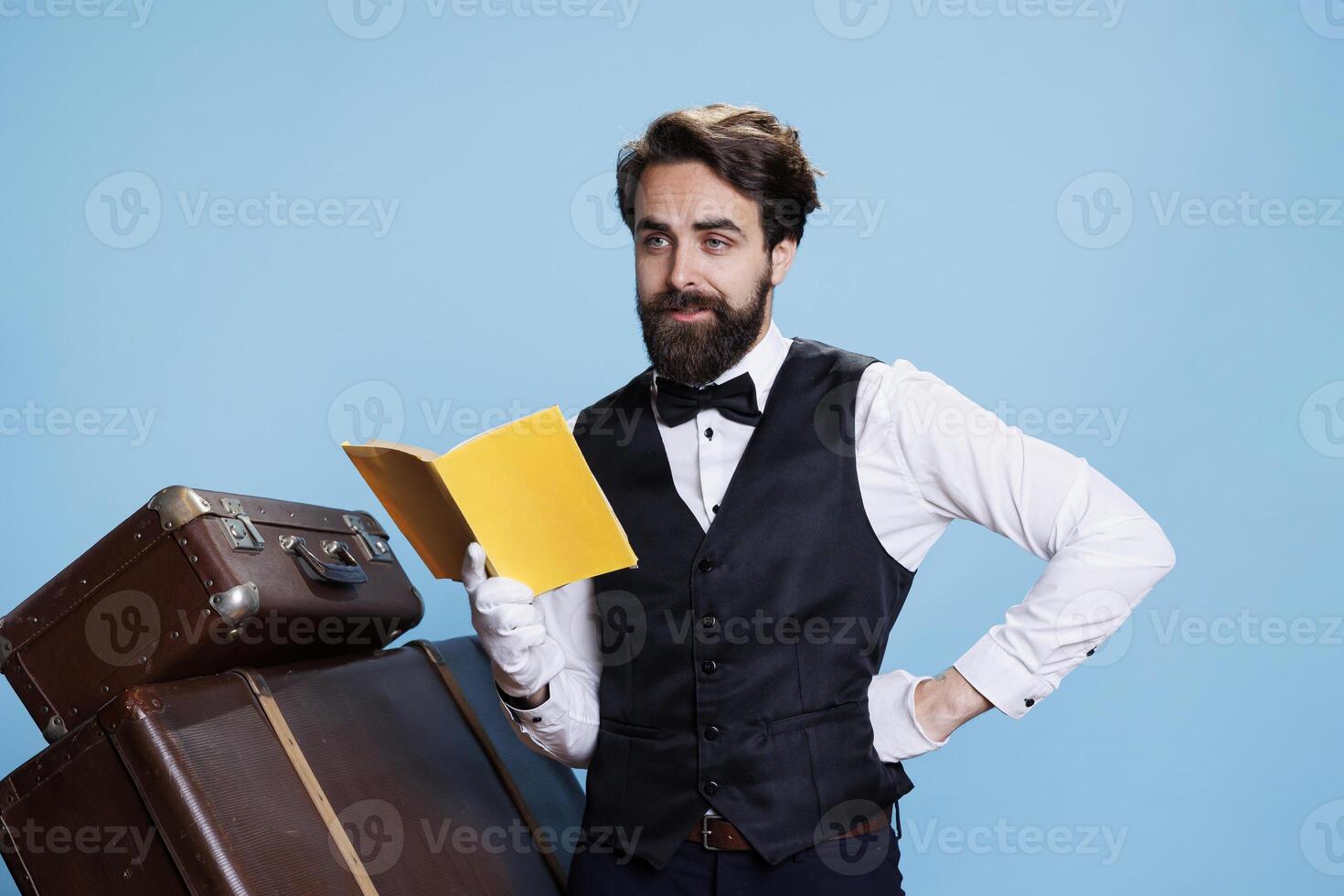 Hotel staff reads fiction book on camera, wearing tie and gloves while he is enjoying literature against blue background. Employee having fun with lecture hobby in studio, novel story. photo