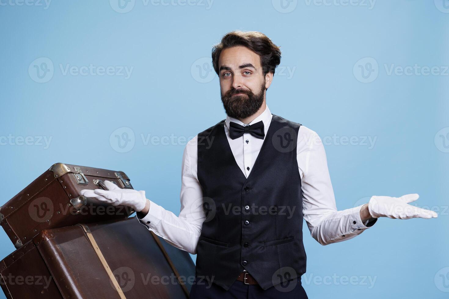Hotel concierge does i dont know gesture, shrugging in front of camera and acting uncertain next to trolley bags. Man bellhop feeling unsure and indicating doubt, unreliable answer. photo