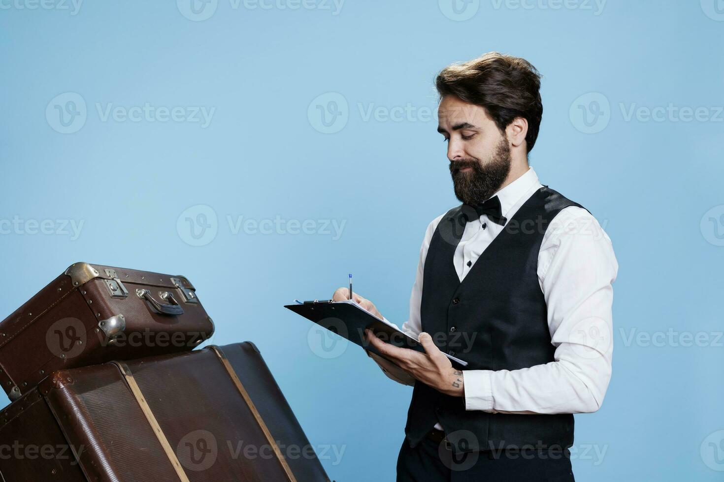 Doorman with tie verifying list on papers, checking names of hotel guests as they arrive. Young man with bellhop occupation takes notes on archive register files to keep evidence on camera. photo