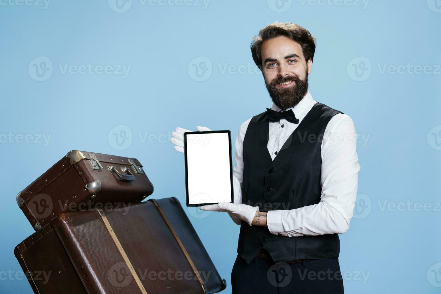 Bellboy presents white blank screen on tablet against blue background, wearing formal suit and gloves next to pile of suitcases. Classy porter showing copyspace template on device. photo