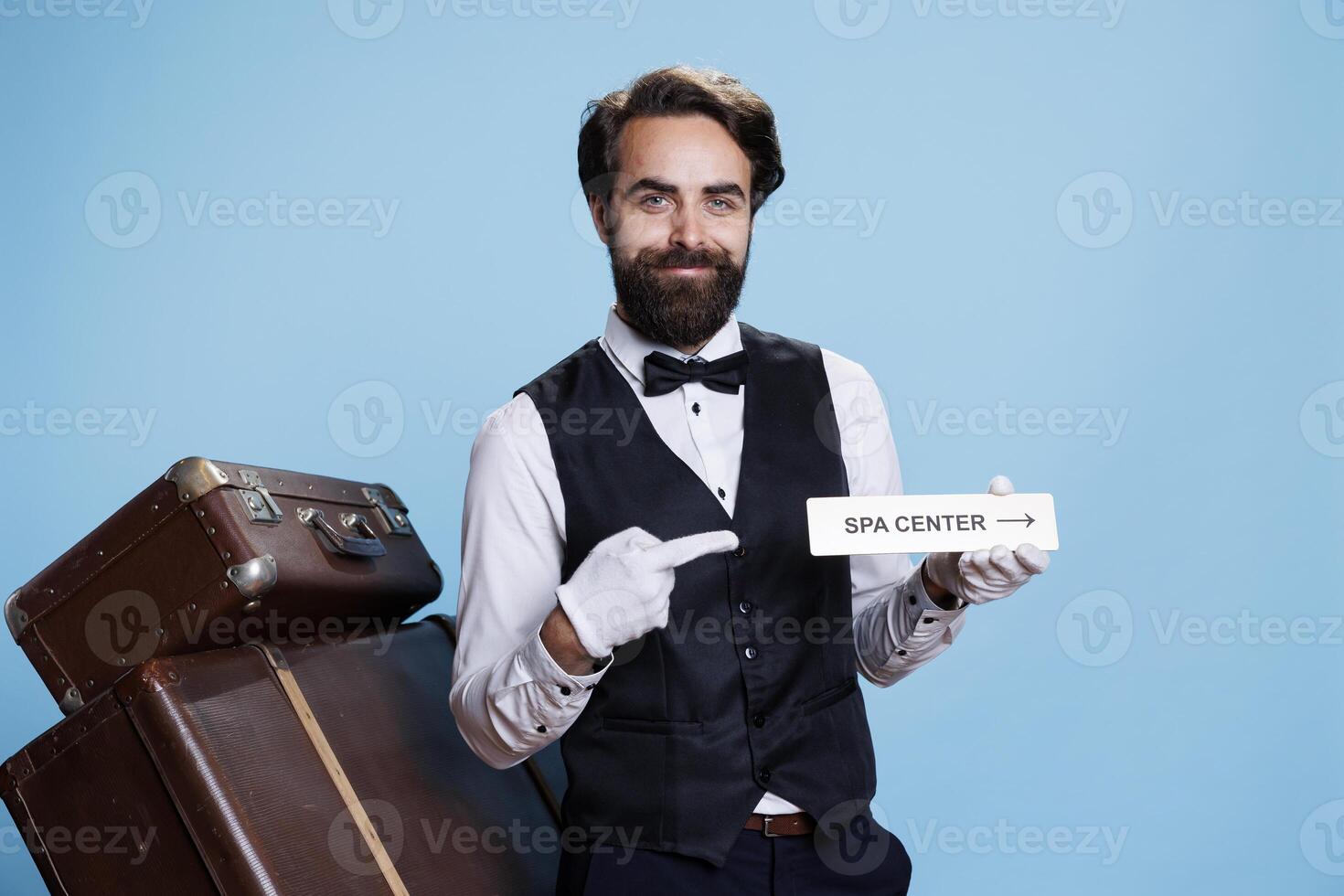 Smiling bellhop poses with spa sign to guide visitors and present different hotel areas, tourism industry. Cheerful professional doorman holding indicator as direction for relaxation area. photo