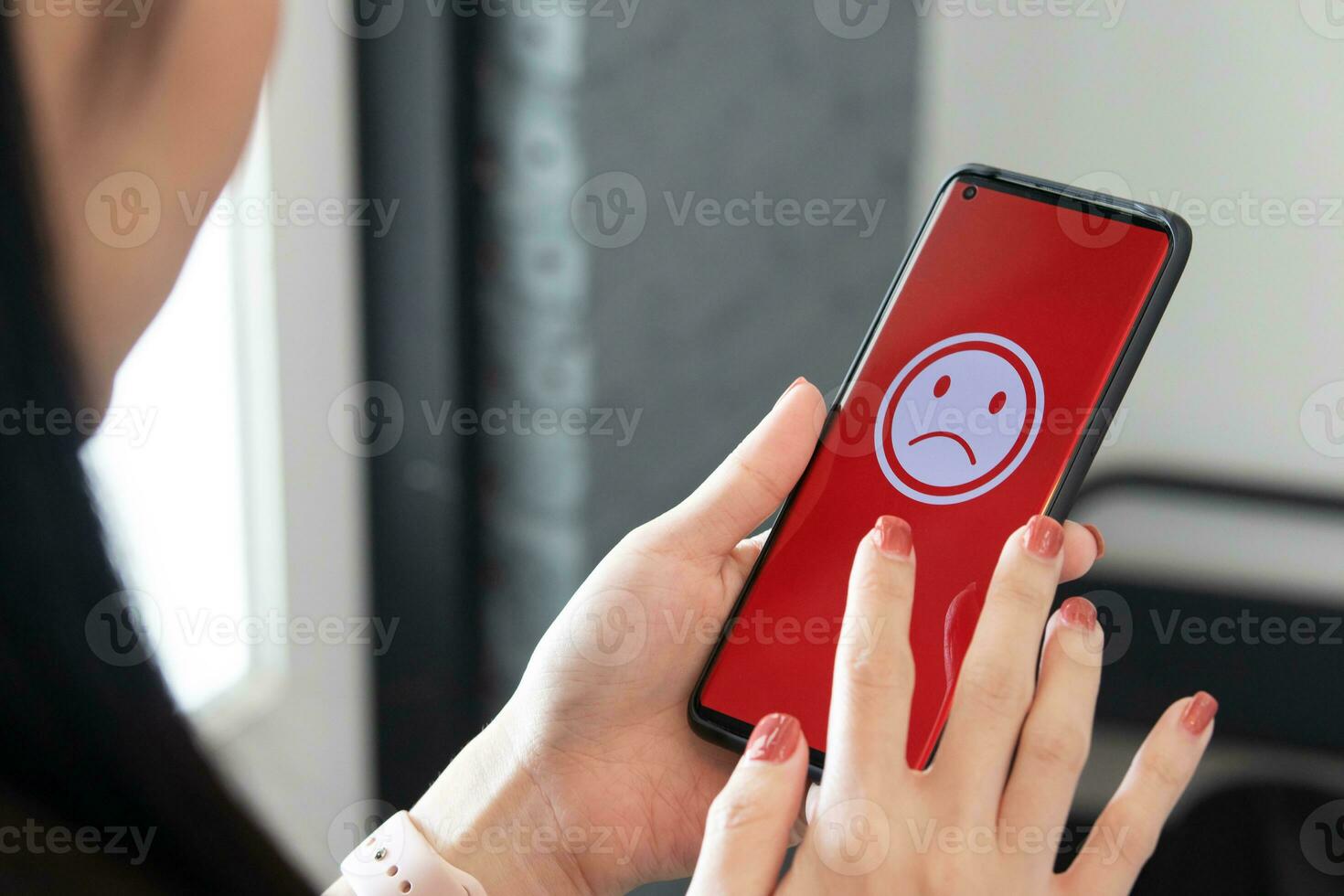 A woman holding a smartphone with a bad feeling emoticon on screen at a restaurant. concept of evaluating customer satisfaction on service levels and recommending that stores should improve. photo