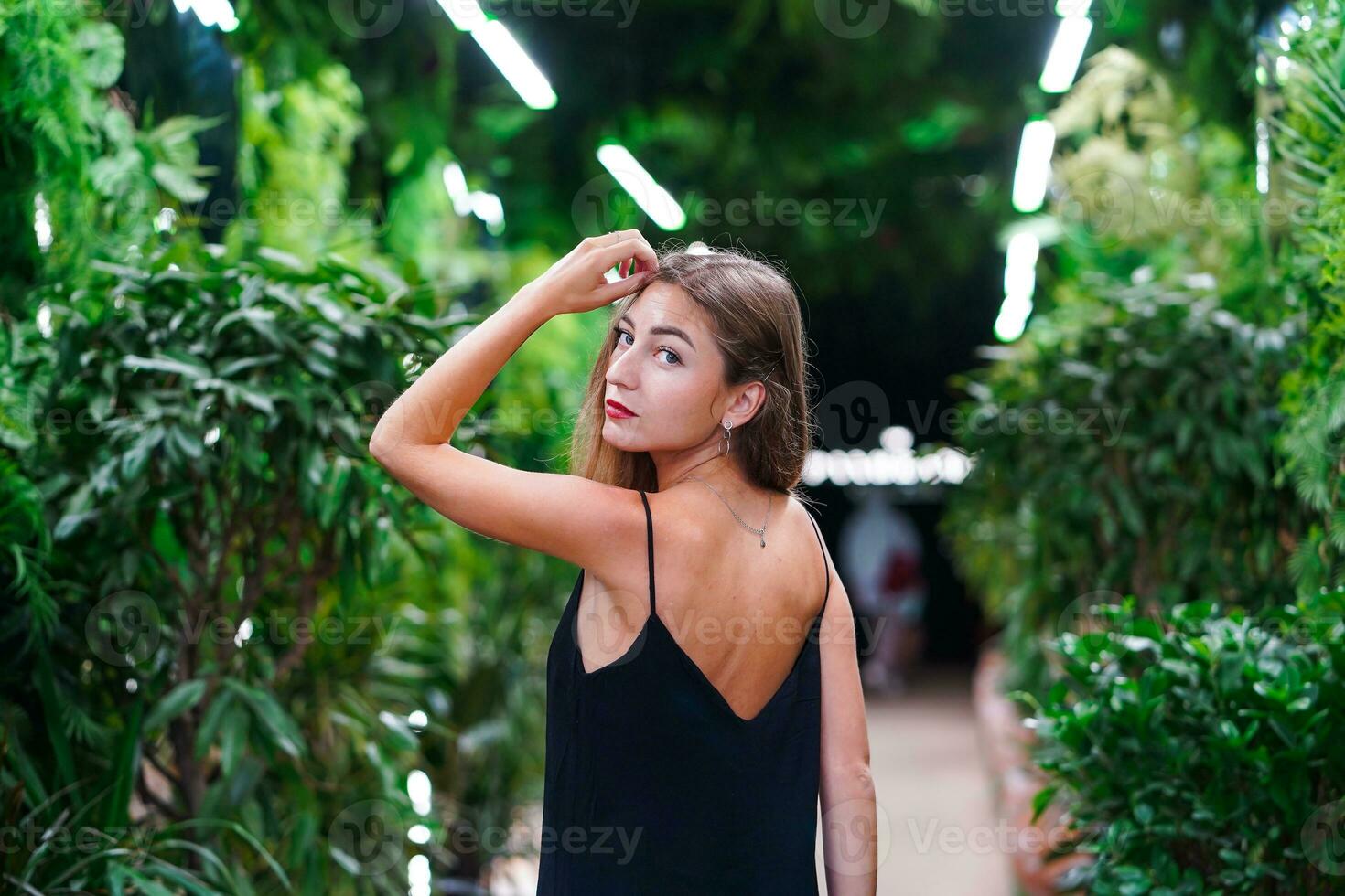 Back view of Young woman wearing black dress with open back standing among green plants. Girl holds hand near her head. Happy young woman walking in tropical garden in black dress, enjoying nature. photo