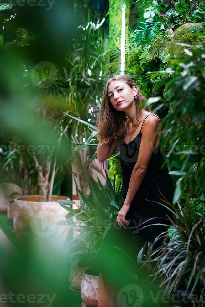 hermosa mujer en pie y mirando directamente a cámara. tropical plantas. joven mujer vistiendo negro vestir dentro invernadero. elegante y elegante moda. retrato de atractivo joven morena mujer. foto