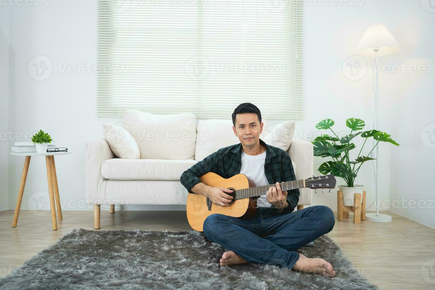 Asian man playing guitar sitting on sofa couch or floor in the living room at home. Asian man writing song while playing guitar at home. Compose song music concept. photo